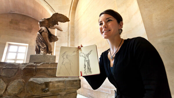 Girl holding sketchbook in front of Winged Victory of Samothrace, Louvre Museum