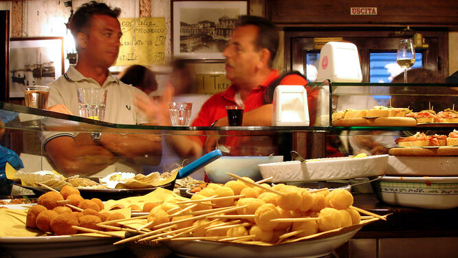 Two men chatting animatedly behind a bar holding plates of fried snacks and other cicchetti, in a characteristic Venetian bar