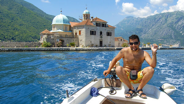 Montenegran man in swim trunks operating a small motorboat in the Bay of Kotor, with a small domed "Our Lady of the Rocks" Church behind him, and steep Montenegran mountains in the background