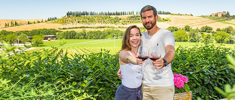 A man and a woman clink their glasses of red wine with Tuscan hills in the background